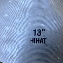 Detailní záběr na kovový činel s koncentrickým žebrováním. Uprostřed je černý nápis 13" HIHAT. Povrch vykazuje drobné škrábance a známky používání.