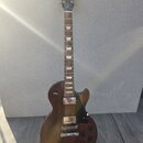Hnědá elektrická kytara s tvarem singlecut a tmavým hracím hředcem. Na hlavě je bílé logo Gibson a nápis Made in USA. Kytara má dva humbuckery, čtyři…