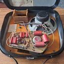 Otevřený černý kufr, v němž se nachází elektronický zesilovač s reproduktorem. Na základní desce jsou vidět různé komponenty, včetně červených válcov…