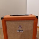 Kytarový kombo zesilovač v oranžovém vinylovém potahu s černými rohovými chráničmi a rukojetí. Přední mřížka je v béžové barvě s dvěma logy: horní ku…