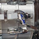 Detailní záběr na chromovaný mechanismus snare thrower na stříbrném bubnu. Mechanismus obsahuje bílý plastový pásek a kovovou páku. Povrch je lesklý,…