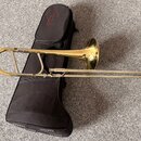 Zlatý mosazný trombon položený vedle černého měkkého pouzdra. Na pouzdře je v horní části červené kulaté logo s nápisem TENOR. Nástroj vykazuje v hor…