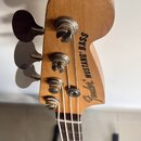 Zadní strana hlavy basové kytary z přírodního dřeva. Na zadní straně je černý nápis Fender MUSTANG BASS. Jsou vidět čtyři kovové ladičky s oválnými k…