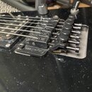Detail černé elektrické kytary s instalovaným černým tremolo mostem. Na základní desce mostu je viditelný bílý nápis Floyd Rose. Povrch kytary vykazu…