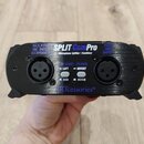Černý kovový přístroj s nápisem SPLIT Com Pro a podtitulem Microphone Splitter / Combiner. Na přední straně jsou dva konektory XLR, přepínače GROUND…