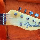 Hlava elektrické kytary z přírodního světlého dřeva s šesti kovovými ladičkami. Na hlavě je černé logo Fender a nápis "STRATOCASTER". V horní části j…