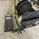 Na fotografii je černý vícekanálový rozvodový box s řadou kombinovaných XLR konektorů a širým D-Sub konektorem na horní straně. Vedle něj je kabel s…