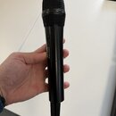 Černý dynamický mikrofon s kovovou mřížkou. Na těle je svisle napsán bílý nápis SENNHEISER a nad ním menší, červený nečitelný text. Na spodní části j…