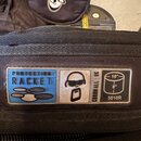 Detail černé textilní brašny s nápisem PROTECTION RACKET na modrém pozadí. Vedle je bílý čtverec s ikonou hlavy v sluchátkách a nápis CORNWALL - UK.…