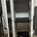 Bílá kovová serverová skříň s otevřenými dveřmi. V interiéru jsou umístěny tři černé police na různých úrovních. Povrch skříně vykazuje běžné známky…