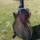 Zadní strana kytary s tmavě fialovým průsvitným lakem a viditelnou strukturou dřeva. Instrument má žlutý lem po obvodu těla a je umístěn na černém st…