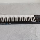 Černý MIDI kontrolér s klaviaturní klávesou. Na přední straně je bílý nápis ALESIS V49 a vpravo se nachází sekce s osmi čtvercovými pady, několika ot…