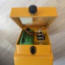 Otevřený žlutý plastový box, v němž je umístěna zelená baterie s logem GP. Z baterie vedou červený a černý vodič k vnitřnímu mechanismu, který obsahu…