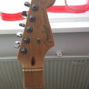 Fotografie zachycuje hlavu elektrické kytary v přírodním dřevě s chromovanými ladičkami. Na hlavě je černý nápis Fender, STRATOCASTER® a menší text C…