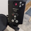 Černý ovládací modul elektronických bicích s červeným digitálním displejem, na kterém svítí číslice 9. Na přední straně je bílý nápis Roland a pod ní…