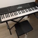 Černý digitální klavír značky YAMAHA na černém skládacím stojanu s přívodní židlí. Nástroj má bílé a černé klávesy, ovládací panel s tlačítky a displ…