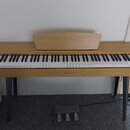 Digitální piano v dřevěném provedení s bílými a černými klávesami. Na středové liště pod klaviaturou je nápis HENRY'S. Nástroj stojí na čtyřech šedýc…