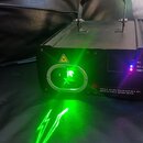 Černý laserový projektor v kovovém provedení s úchytem na horní straně. Na přední části je žlutý varovný trojúhelník a nápis: Visible or invisible ra…