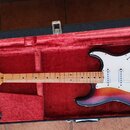 Elektrická kytara s telyem v červeno-žlutém sunburst provedení a bílým pickguardem, uložená v černém kufru s červeným vnitřkem. Na hlavě hředu je vid…