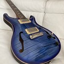 Modrá semiakustická kytara s vyznačeným slibovnicovým vzorem dřeva a dvěma f-otvory. Nástroj má dva humbuckery s krémovými rámečky, stříbrný most a t…