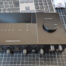 Černý audio interface s nápisem KOMPLETE AUDIO 6 na přední straně. Horní panel obsahuje kontrolky s popisky IN 1, IN 2, IN 3, IN 4, OUT 1/2 a USB, 48…