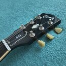 Černá hlava elektrické kytary s bílým logem Gibson a nápisem SG. Na hluboce černém lakovaném povrchu je vidět bílý ornament ve tvaru ptačího rozpětí.…