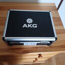 Černý transportní kufr s hliníkovým rámem a kovovými zapínacími zámky. Uprostřed víka je bílé logo a nápis AKG®. Na přední straně kufru je nalepená b…