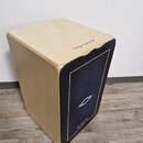 Dřevěný cajon s přírodním povrchem a černým předním panelem. V horní části je nápis „Hand Artisan Edition“. Na černém povrchu je viditelné bílé logo…