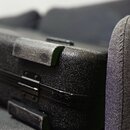 Detailní záběr na hranu černého, texturovaného kufru s tvrdým povrchem. Jsou vidět kovové panty a dvě ochranné gumové nebo plastové nárazníky v horní…