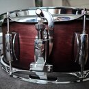 Detailní záběr na snare drum s tmavě hnědým dřevěným korpusem a chromovanými obručmi. Na chromovaném mechanismu napnutí je vyryto logo "Gretsch". Nás…