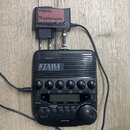 Černý elektronický metronom TAMA RHYTHM WATCH RW200 s ovládacími tlačítky a otočným knoflíkem. K přístroji je připojen černý napájecí adaptér, na kte…