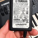 Černý síťový adaptér značky YAMAHA. Na bílém štítku je uveden model PA-150B, vstupní napětí 100-240V~ 50/60Hz 0.5A a výstupní napětí 12,0V 1.5A 18.0W…