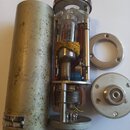 Fotografie zobrazuje rozebraný kovový mikrofon stříbrné barvy. Vnitřní mechanismus obsahuje elektronickou lampu a cívku, přičemž jedna část je omotan…