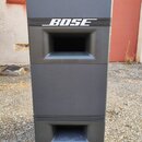 Černý lineární reproduktor v plastovém provedení s kovovými rohy. Na horní části je bílý nápis BOSE umístěný mezi dvěma vodorovnými linkami. Povrch j…