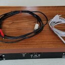 Na dřevěném podstavci s imitací orechu leží černý přístroj a dva kabely (černý audio kabel s RCA konektory a šedý napájecí kabel). Na zadní straně če…