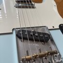 Detail elektrické kytary v modré barvě s bílým pickguardem. Na kovové základně snímače je vyražen nápis FENDER PAT. PEND. Snímač je černý a pod ním s…