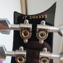 Detail zadní strany černé hlavy kytary s kovovými ladicími mechanismy. V horní části je žlutým nápisem uvedeno číslo 17 244383. Povrch laku je hladký…