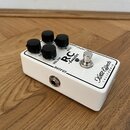 Bílý kytarový efekt pedál s kovovým šasi a čtyřmi černými otočnými knoflíky. Na horní straně jsou nápisy VOLUME, RC booster a značka Xotic Effects CA…
