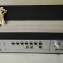 Černý elektronický přístroj s hliníkovým chladičem v horní části a šedým zadním panelem. Na panelu jsou umístěny vstupy, přepínače a nápisy: 220V-1A/…