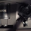 Černý mikrofon s nápisem RØDE umístěný v originální pěnové výplni. Vedle něj je srolovaný černý kabel USB a držák mikrofonu. Všechny komponenty vypad…