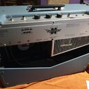 Modrý kytarový zesilovač s kovovým zadním panelem. Na panelu je vidět logo a název modelu Vaporizer, značka Musical Instruments Corp. a označení CE.…