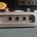 Zadní strana šedého elektronického zařízení s třemi jackovými konektory a jedním napájecím vstupem. Nad konektory jsou nápisy INPUT, OUTPUT (PHONES)…