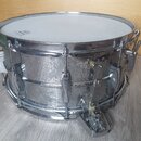 Stříbrný kovový buben s bílou blánou. Povrch korpusu vykazuje výrazné opotřebení, oxidaci a odsloupání chromového povrchu. Na bláně je vidět nečiteln…