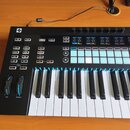 Černý MIDI kontrolér s klaviaturní klávesou. Na horním panelu jsou umístěny knofíky, tlačítka a mřížka bílých podsvícených padů. V levém horním rohu…