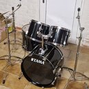 Černá bicí souprava značky TAMA. Sestava obsahuje basový buben s logem TAMA na přední bláně, dva tom-tomy a jeden floor tom. Všechny bubny mají chrom…