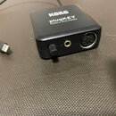 Černý obdélníkový adaptér s kabelem a konektorem Lightning. Na horní straně je bílý nápis KORG plugKEY MOBILE MIDI/AUDIO. Na boční straně jsou umístě…