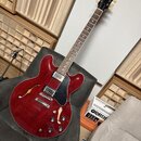 Polodupatá elektrická kytara v červené barvě s černým pickguardem. Na hlavě hmatníku je viditelné logo Gibson. Kytara má dva humbuckery, tři ovládací…