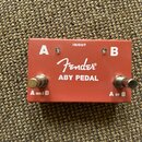 Červený kovový kytarový pedál s nápisem Fender ABY PEDAL. Na horní straně jsou bílá písmena A a B, nápis IN/OUT a dva kovové spínače s popisky A and…