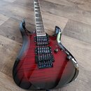 Elektrická kytara s červeným gradientním lakem a viditelnou strukturou dřeva. Nástroj má dva humbuckery, hmatník s vložkami a černý most. Na kytaru j…