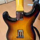 Zadní strana elektrické kytary s tělem v provedení sunburst. Viditelný je krk z jasného dřeva a kovový spojovací plech u základny hmatníku. V dolní č…